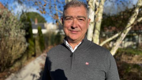 A man is standing outdoors, looking at the camera and smiling. He is wearing a grey jumper. There are trees and bushes with a path in the background. The sky is blue. 