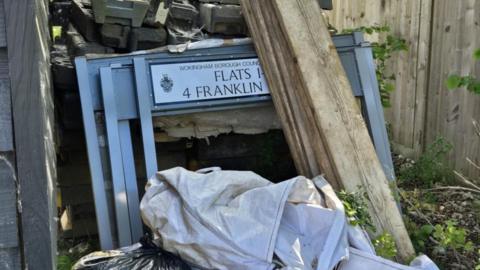 A pile of fly-tipped rubbish, including wooden planks and a white tarpaulin. There are also three road signs, with the one at the top of the pile clearly slowing the Wokingham Borough Council logo.