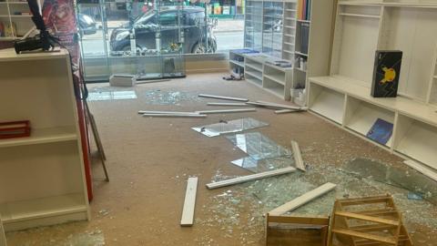 Smashed glass and broken shelving covers the floor of Trove indoor market in Winton. The shop window looks out on to a street with a parked car outside.