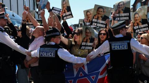 Three police officers stand in front of a group of protesters holding signs reading: "Keir Starmer Jew Harmer". The placards also depict a picture of former Labour leader Jeremy Corbyn holding a mask of Keir Starmer in front of his face. 