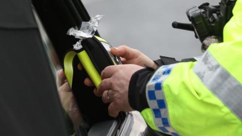 A police officer in a high vis jacket holds a breathalyser up to the window of a car.