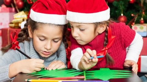 Two young girls wearing red and white Santa hats are colouring green paper Christmas trees in front of a large Christmas tree. Both have pencils in their hands and one is looking at the other's design.