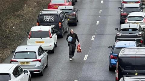 A man in a black hoodie walks down a stretch of road between queues of traffic. He has a large bag of crisps in his hand and bottled water. 
