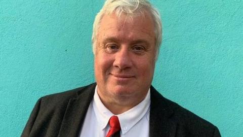 John Wannenburgh, who had short blond hair and is wearing a black jacket over a white shirt and red and black tie.
