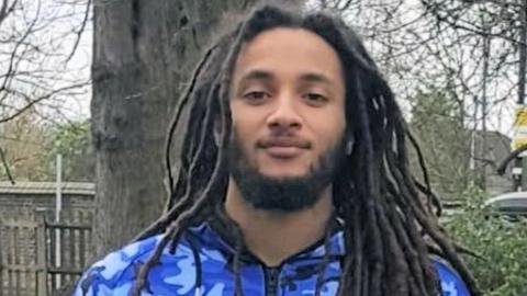 Nimroy Hendricks, who has a short beard and long dreadlocked hair and is wearing a tracksuit top with a blue camouflage design. He is stood in front of a tree, looking gently at the camera.