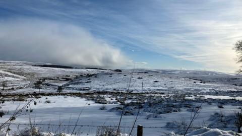 Snow covering the hills on Dartmoor, with blue and cloudy skies seen afar.