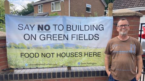 A man with grey hair and a grey beard, wearing a brown T-shirt, stands in front of a brick wall, which has a large campaign banner attached to it, saying Food Not Houses Here