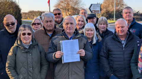 A group of men and women standing by a road outdoors. At the centre is council leader Dave Blackwell, who is holding a letter in a plastic sleeve.