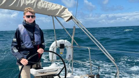 The image shows a person steering a sailboat through the ocean. They're wearing a life jacket and casual clothes. The background includes a wide view of the ocean with waves and a partly cloudy sky.