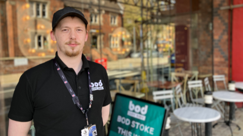 A man in a black polo shirt and black cap, with a lanyard around his neck. He is standing in front of large glass windows in which the reflection of buildings opposite can be seen.