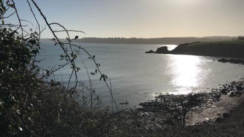 A cove is seen with the sun shimmering on the sea and a rock formation in the background 
