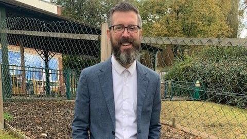 A man with short hair, glasses and a long beard, stands in a garden behind a school with a fence just behind him. He is wearing a grey suit jacket and a white shirt.