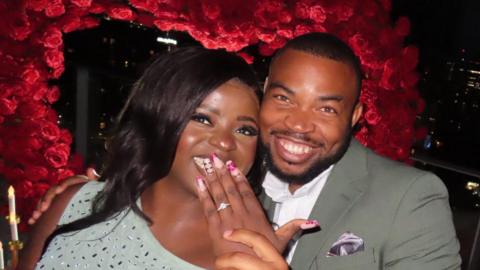 Malaika and Fred grin as she raises her hand with an engagement ring to her smile. They lean into each other in front of a large heart-shaped stand of red roses with some candle lights on both sides at night. She is wearing a sparkly grey dress and he is wearing a grey suit and white shirt.