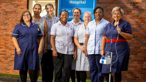 A group of eight women wearing nursing uniforms, one on the right of the picture is holding a tube connected to a patient pump