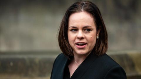 Head-and-shoulders shot of Kate Forbes, who has shoulder-length brown hair, looking off to her left. She is wearing a black blazer-style jacket and standing outside a grey church building. Her mouth is open as if she is talking to someone. 