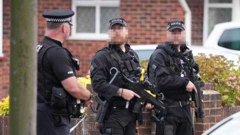 Three armed police at the scene of an incident in Lindum Road, Worthing. Two of the officers' faces are blurred out. All three are wearing black uniform and police caps. Two guns are visible in the image.