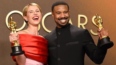 Jessie Buckley and Michael B Jordan standing together, smiling and holding up their Oscars