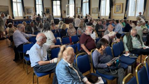 A room full of people sitting on a chairs in a row at a public meeting.