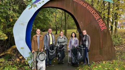 Stewart Turnbull (second left) with four volunteers at an entrance to Gateshead Riverside Park. They are each carrying litter pickers and rubbish bags. They are standing in front of a large sign for the park. Behind them are trees and a path.