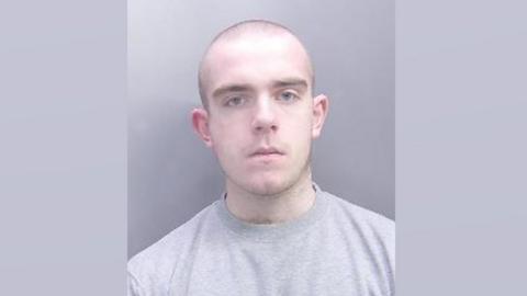 A custody image of Noah Hardingham. He has a shaved head and is wearing a grey T-shirt and sitting against a grey background