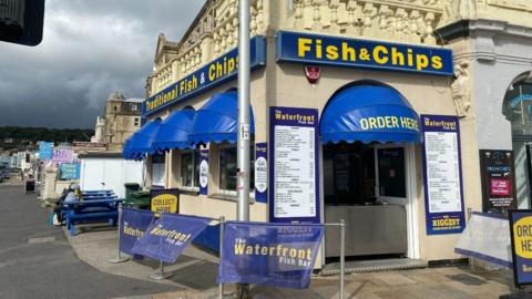 A fish and chips shop with blue signs. There are blue picnic benches next to the shop.