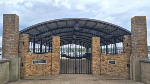 The yellow brick entrance to Rochester Pier. The gap in the walls are blocked by a black metal gate, while the pier behind is sheltered by a curved roof.