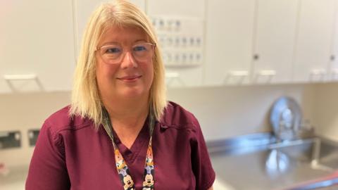 A woman with long blonde hair, glasses, a Disney-themed lanyard and a burgundy top, stands in a room in a hospital and smiles.