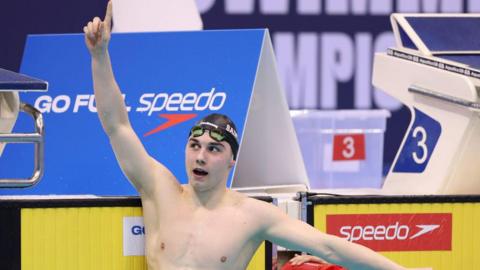 Will Ellard in the pool, wearing goggles and a swimming cap, at the end of a race with his hand and index finger pointed to the sky. 