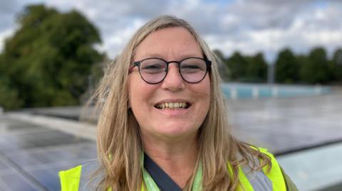 A woman wearing round black framed glasses smiles toward the camera, she is wearing a yellow high-vis jacket and green blouse, behind her are trees and a building rooftop with solar panels.