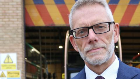 Darren Roe wearing a blue suit, a blue shirt and blue tie. He has black square-framed glasses and is looking directly at the camera. He has white hair and a white beard. 