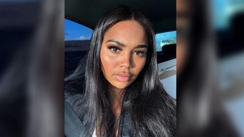 A woman with long straight dark hair sits in a car, wearing a black jacket and looking directly at the camera. Sunlight highlights her face, with a seatbelt visible across her chest and a blue sky outside the window.