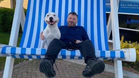 Paul Scott sits on an oversized, novelty blue and white striped deckchair, with a  white bulldog at his side. He is smiling and is wearing a blue top, black trousers and trainers.