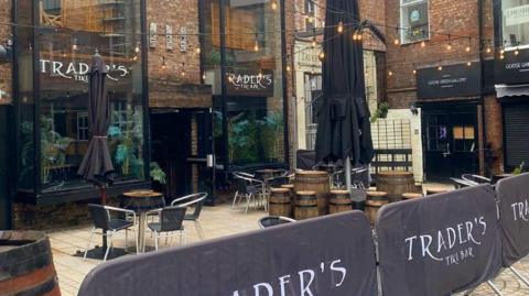 A brick building in a square has signs out the front that read 'Trader's Tiki Bar'. It has tables and chairs out the front. 