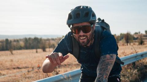 Mitch Hutchcraft cycling during the challenge. He is smiling at the camera and is holding one arm out in front of him. He is smiling and it wearing a helmet and sunglasses. He has a backpack on.