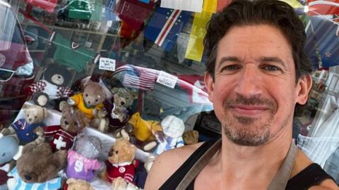 A man with dark hair wearing a dark vest takes a selfie in front of a shop window display that is filled with teddy bears in knitted football shirts.
