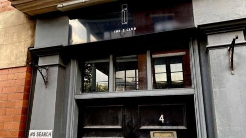 The door of The I Club in Nottingham. The doors are black with glass panels above. A sign above the door says The I Club with a logo above it. 