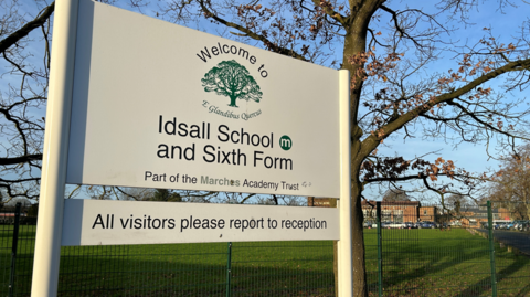 A white sign with Idsall School and Sixth form and an image of a tree on it and a green playing field behind it