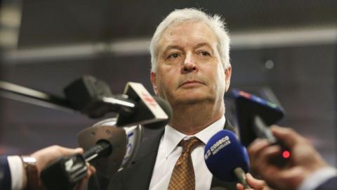 An image showing Michael Rousseau surrounded by media microphones at a scrum in Montreal, pictured in 2021. He has short white hair, and is wearing a suit and copper patterned tie.