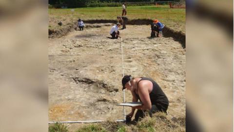 Six people measuring an archaeology trench on a sunny day