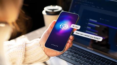 A hand holds a phone showing an image of an An AI chatbot. There is a laptop in the background. 