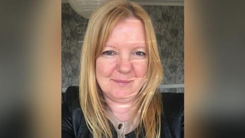 Maxine Hickman, with long blonde hair, wears a black leather jacket and a cream top underneath with gold buttons. She is smiling. She is in a room in a home decorated with grey flowery wallpaper.