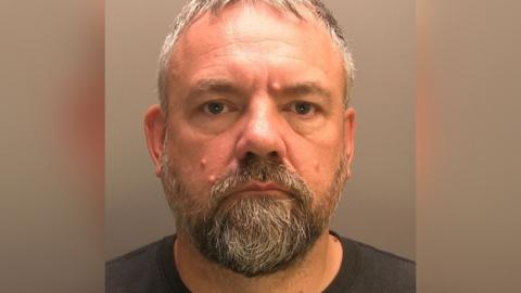 Brett Peterson has short grey hair, a beard and moustache and looks into the camera lense. He is wearing a black T-shirt. 