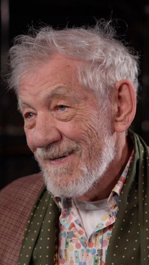 Sir Ian McKellen looking off camera. He is smiling and has grey hair and a beard. He is wearing a patterned shirt and scarf with a brown checked jacket.