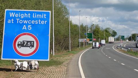 A large blue traffic sign weighed down by sandbags. It reads 'Weight limit at Towcester A5' and shows a red and white symbol of an HGV with '7.5 tonnes' on it. The sign is beside a dual carriageway where light traffic is shown moving freely beneath leaden skies.