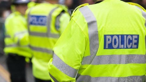 Three police officers standing outside with their backs to the camera. They are all wearing yellow fluorescent jackets with grey stripes along the back and arms. On the back of each jacket is a blue rectangle with the word, POLICE inside.
