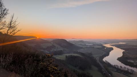 Orange sunrise glow appearing over hilltops in the distance, with a winding river and field and trees also in the shot. 