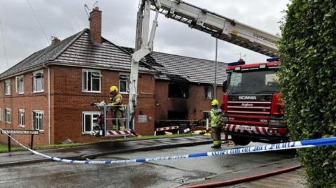 Police tape is cordoning off a road. There is a fire engine with an aerial platform parked next to a block of two-storey flats. A section of the flats is black with smoke damage and there are roof tiles missing. There are two fire personnel, one in the aerial lift, and one standing by the fire engine
