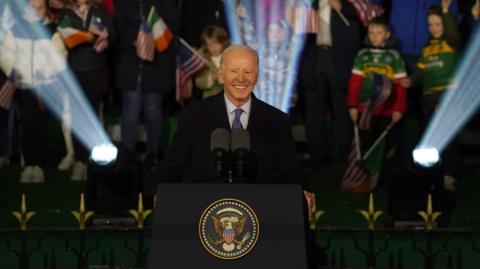 A man with white hair smiles. He is standing at a podium with an American eagle design on the front. He is wearing a suit. Behind him is a crowd, with a mixture of Irish and American flags.