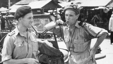 Captain Leonard Rosoman and BBC Engineer Carl Carlyon pictured chatting while leaning on a jeep in Hong Kong. Both are wearing military uniforms and Carlyon is smoking on a pipe.