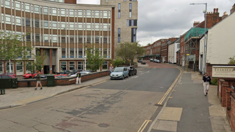 A city centre road, with town houses to the right and an office block to the left, extends away from the camera. It is cloudy above.